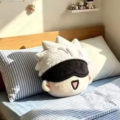 A round plush pillow with a cartoon face wearing a black sleep mask, resting on a bed with blue and white striped bedding and pillows in a sunlit bedroom.